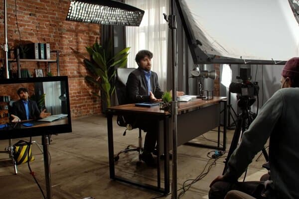 A movie set where an actor is sitting on a desk surrounded by filming equipment and crew